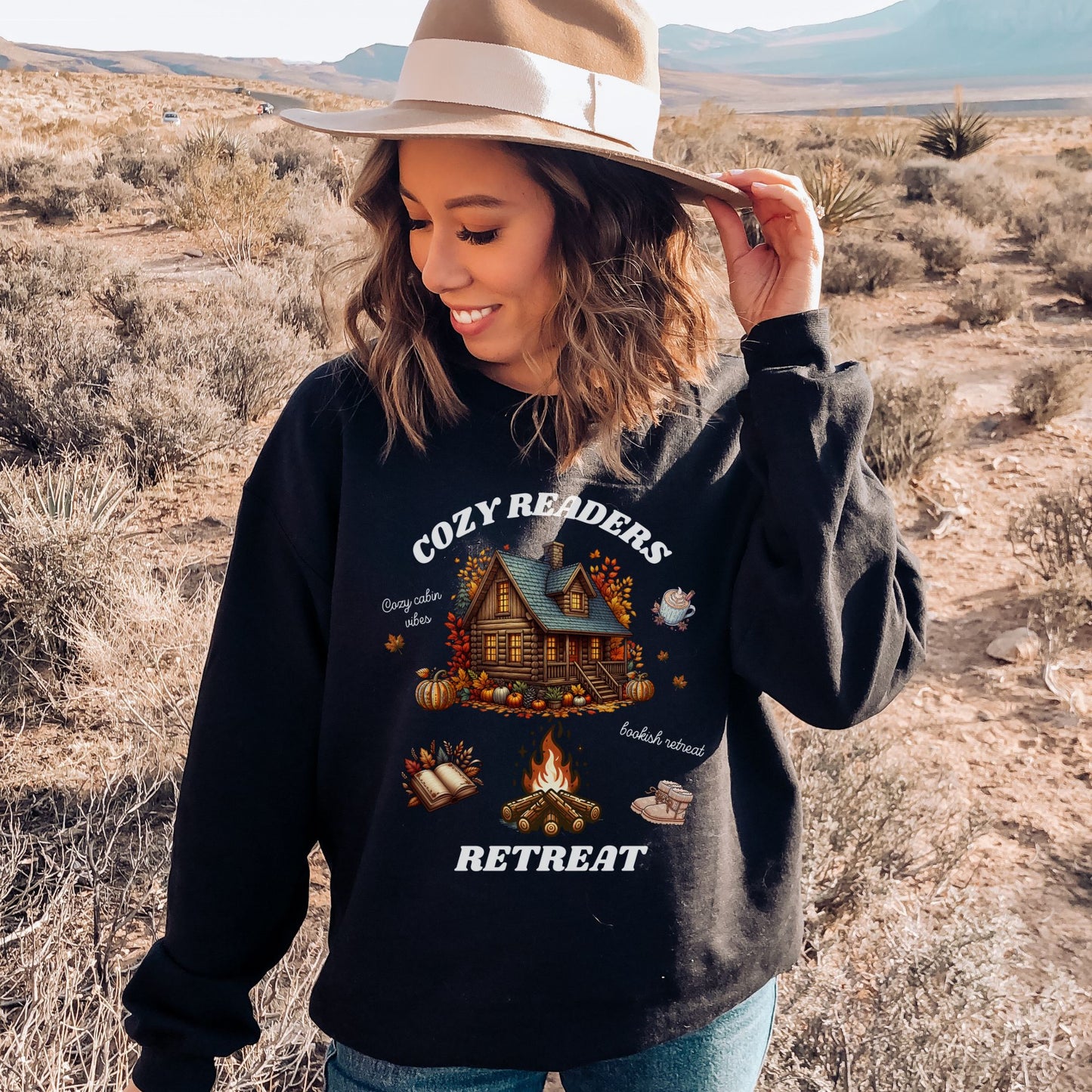 Woman wearing a black Sweatshirt with 'Cozy Readers Retreat' design including a cozy cabin, pumpkin spice latte, cute slippers, campfire, pumpkins and a book. Starlit Prose Bookish Merch Australia