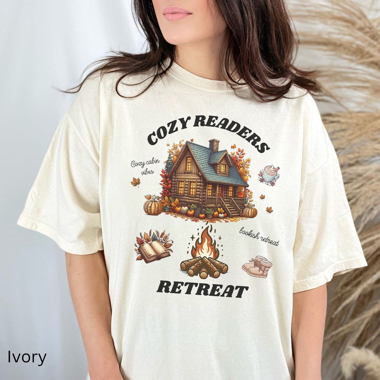 Woman wearing an Ivory Comfort Colors Tee with 'Cozy Readers Retreat' design including a cozy cabin, pumpkin spice latte, cute slippers, campfire, pumpkins and a book. Starlit Prose Bookish Merch Australia