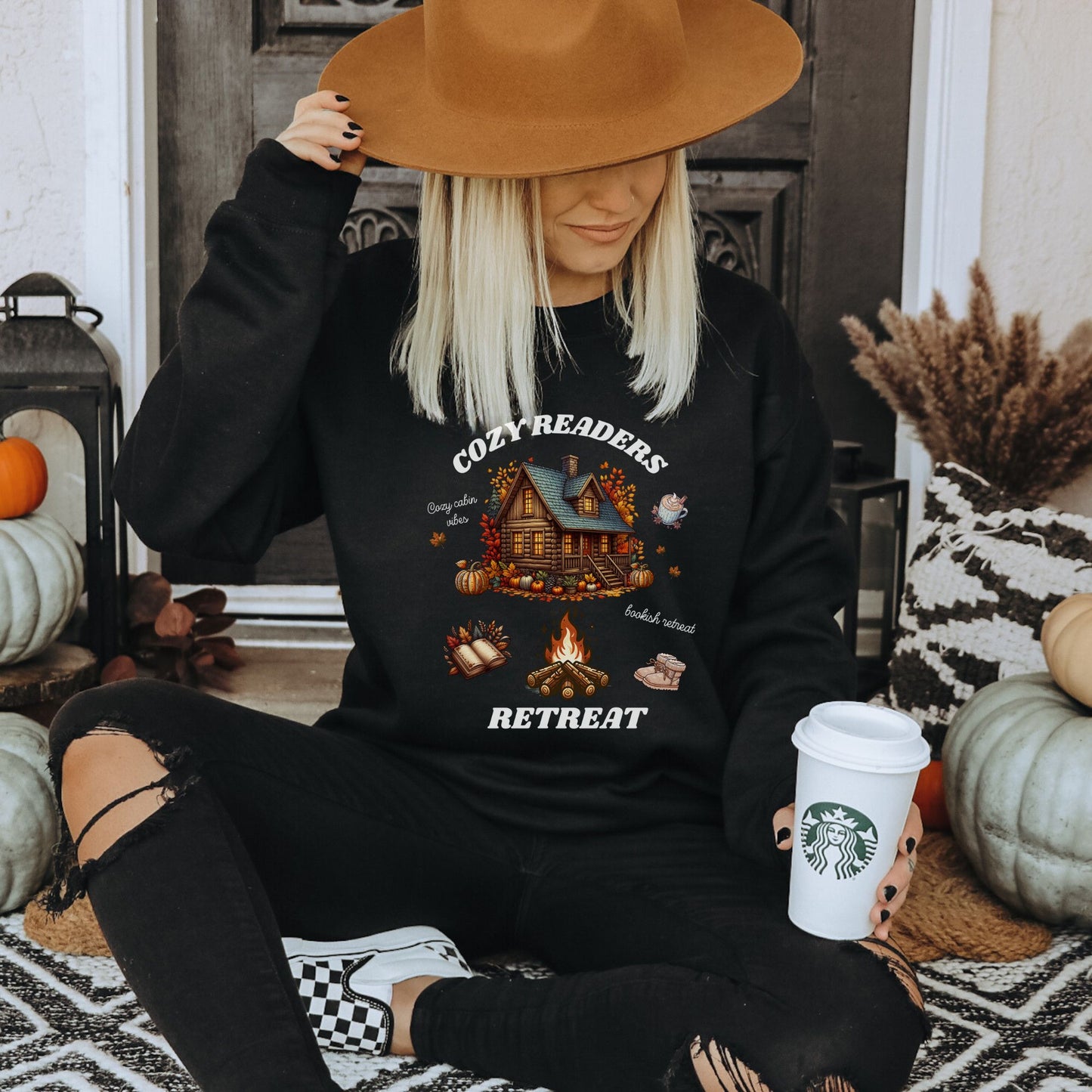 Woman wearing a black Sweatshirt with 'Cozy Readers Retreat' design including a cozy cabin, pumpkin spice latte, cute slippers, campfire, pumpkins and a book. Starlit Prose Bookish Merch Australia
