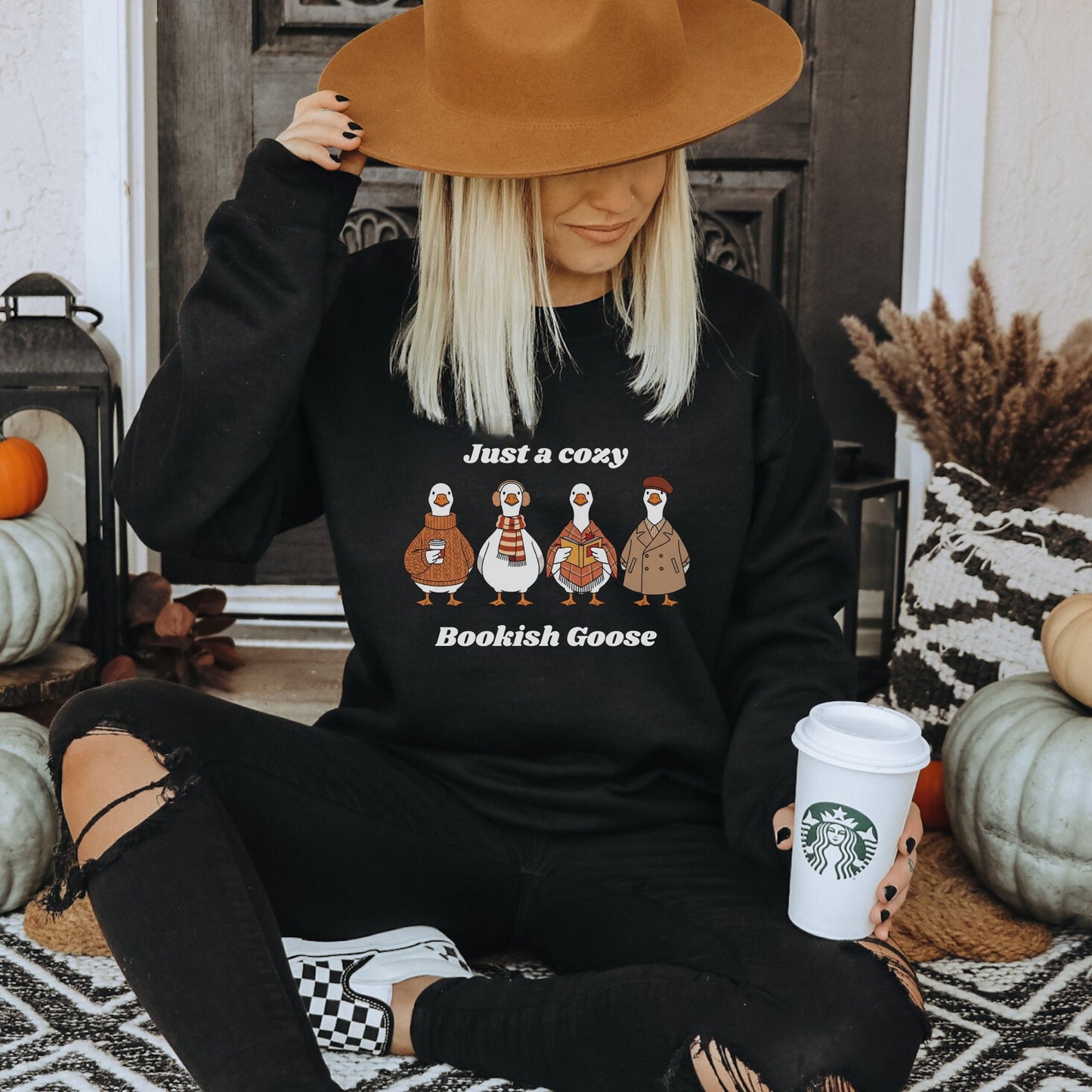 Person wearing a black sweatshirt with cartoon geese that says "Just a Cozy Bookish Goose" and holding a Starbucks cup, sitting on a patterned rug. Starlit Prose Bookish Merch Australia