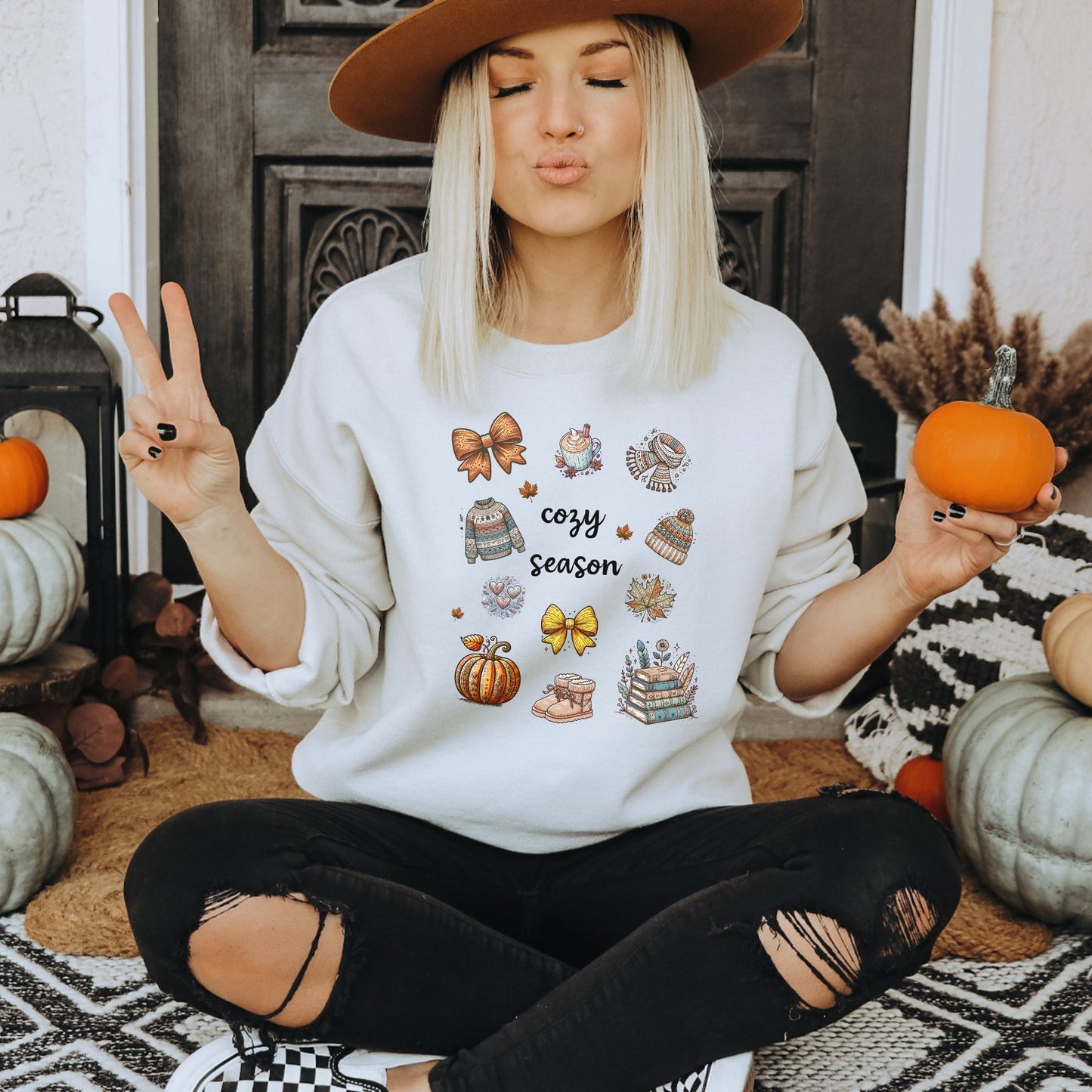 Woman wearing a white sweatshirt with Fall-themed graphics and text, including a Pumpkin Spice Latte, scarf, beanie, stack of books, sweater & pumpkins sitting in a decorated room with pumpkins. Starlit Prose Bookish Merch Australia