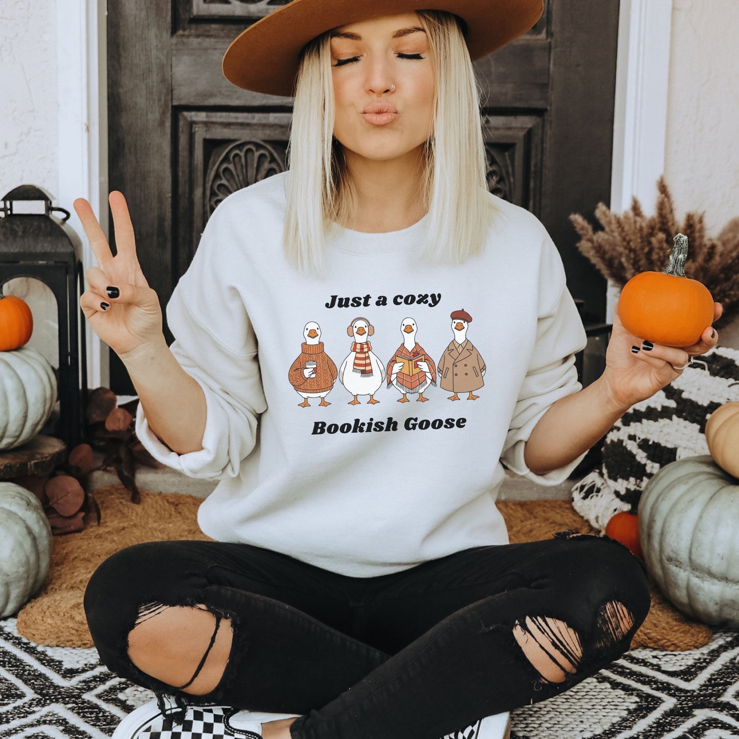 Woman wearing a white sweatshirt with 'Bookish Goose' design, sitting in a cozy indoor setting with pumpkins.