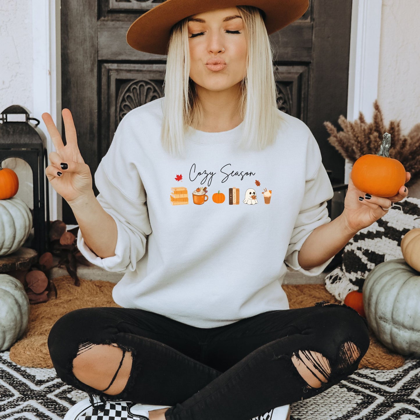 A Woman wearing a white sweatshirt that says "Cozy Season", it includes cute orange Fall Autumn graphics like a stack of books, pumpkin spice latte, pumpkins, a ghost and leaves. Starlit Prose Bookish Merch Australia