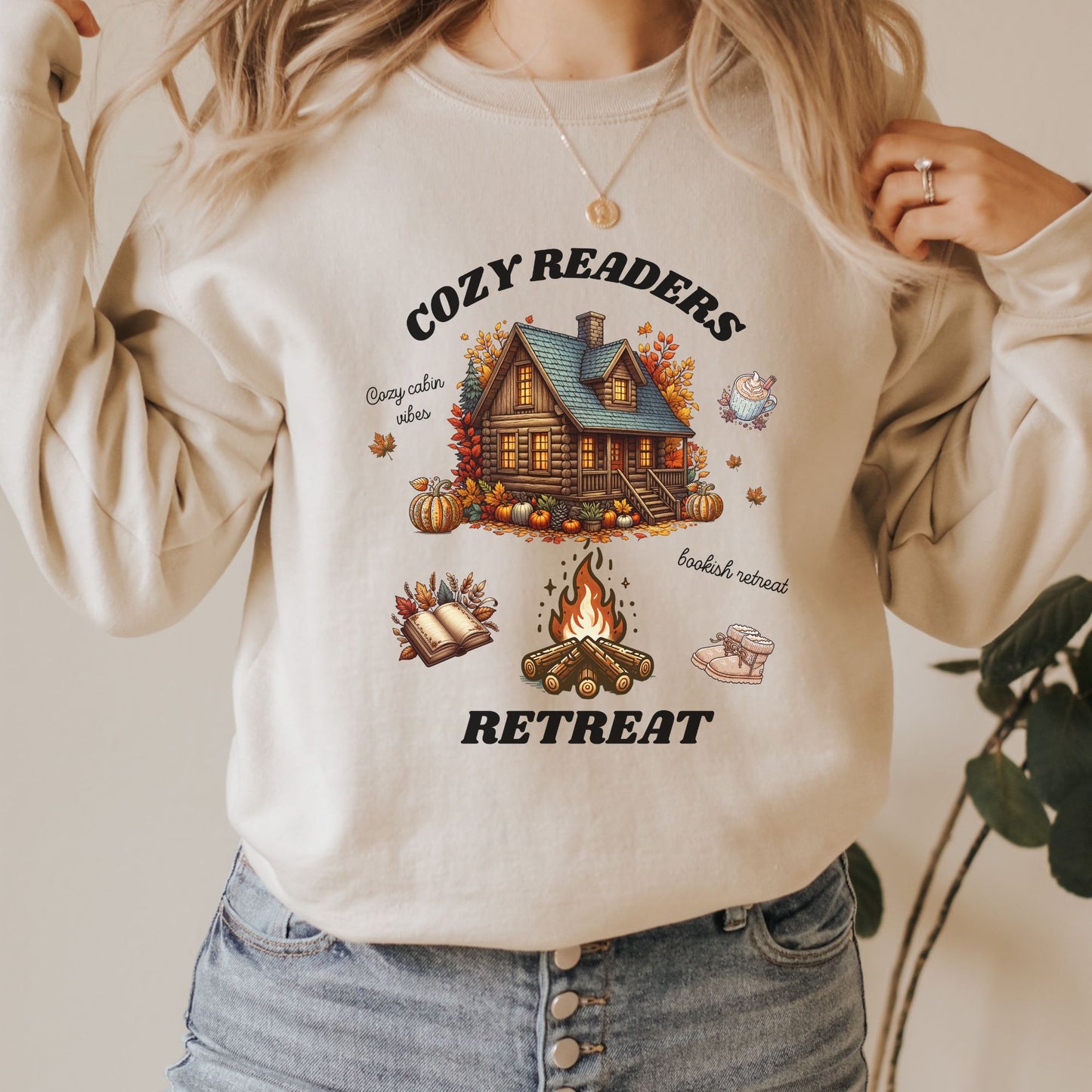 Woman wearing a Sand Sweatshirt with 'Cozy Readers Retreat' design including a cozy cabin, pumpkin spice latte, cute slippers, campfire, pumpkins and a book. Starlit Prose Bookish Merch Australia