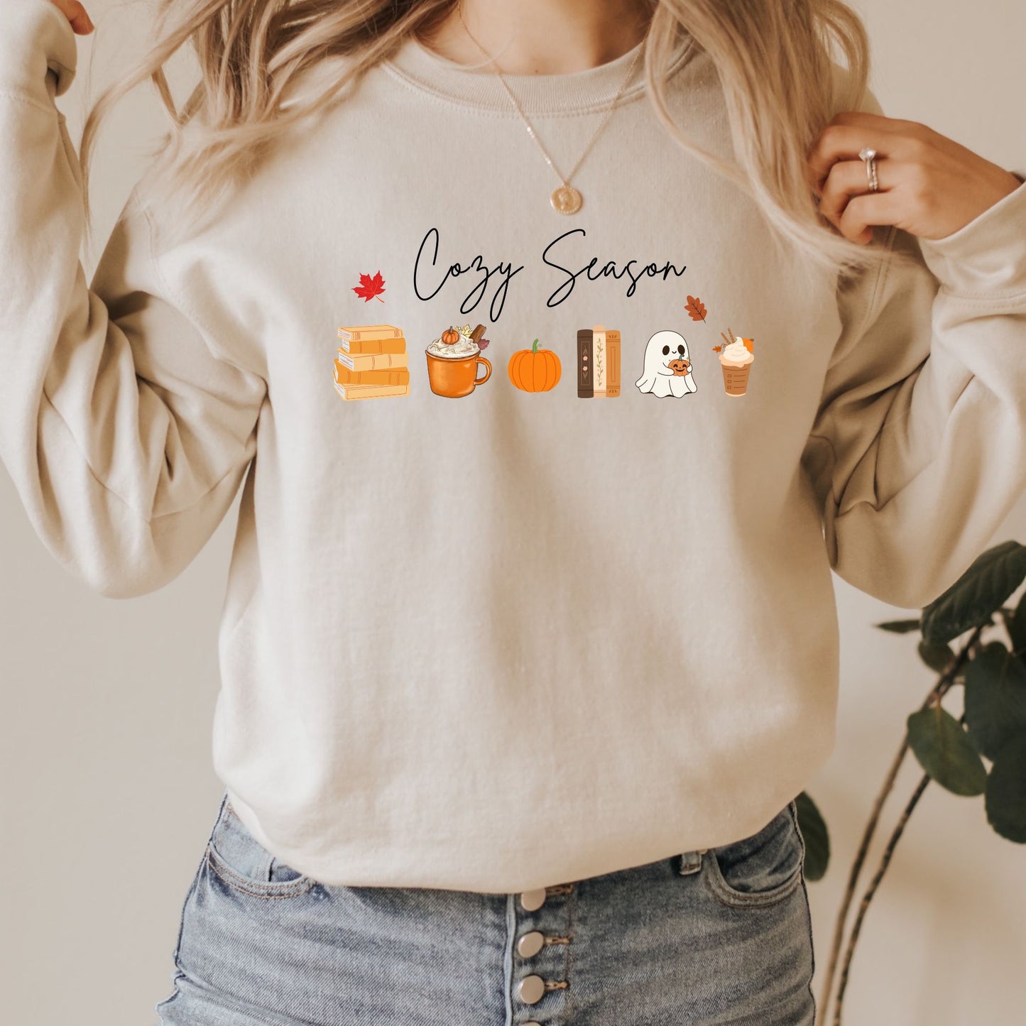 A Woman wearing a Sand sweatshirt that says "Cozy Season", it includes cute orange Fall Autumn graphics like a stack of books, pumpkin spice latte, pumpkins, a ghost and leaves. Starlit Prose Bookish Merch Australia