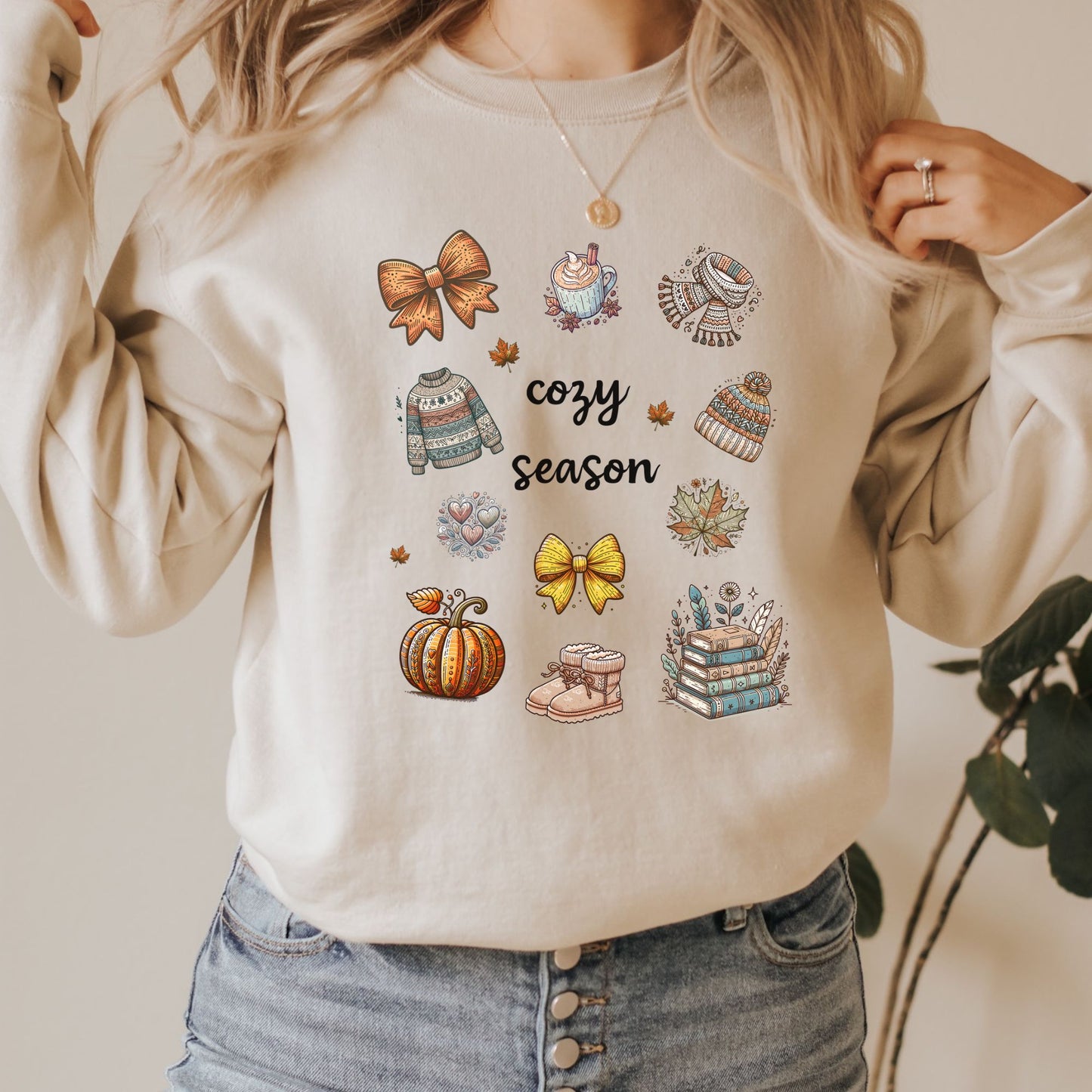 Woman wearing a sand sweatshirt with Fall-themed graphics and text, including a Pumpkin Spice Latte, scarf, beanie, stack of books, sweater & pumpkins sitting in a decorated room with pumpkins. Starlit Prose Bookish Merch Australia