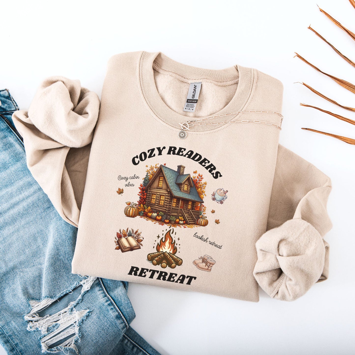 Flat lay of a Sand Sweatshirt with 'Cozy Readers Retreat' design including a cozy cabin, pumpkin spice latte, cute slippers, campfire, pumpkins and a book. Starlit Prose Bookish Merch Australia