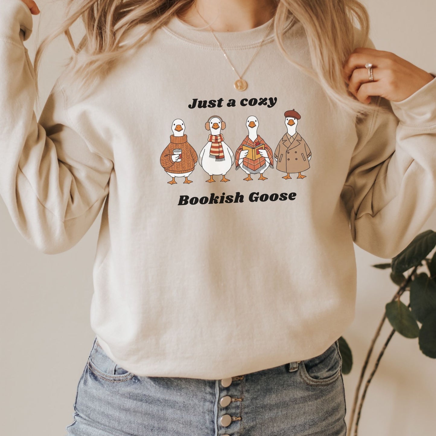Sand colour sweatshirt with cartoon geese and text that says "Just a Cozy Bookish Goose" worn by a person, with a neutral background. Starlit Prose Bookish Merch Australia