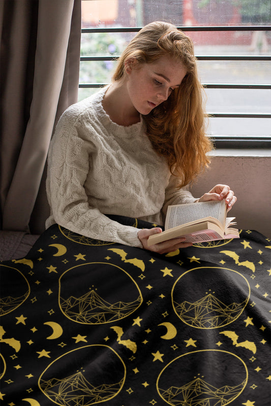 A person sitting on a couch reading a book, with a black and gold geometric patterned throw blanket on the couch.