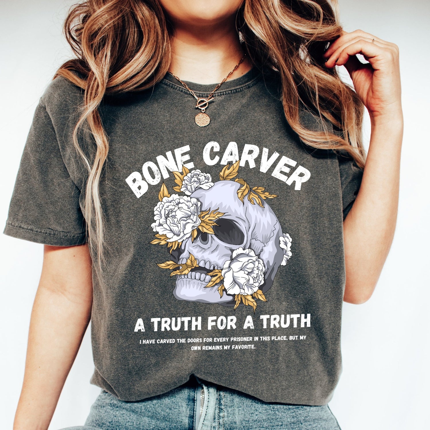 Woman wearing a pepper black comfort colors tshirt with a graphic of a skull with roses. The words say - Bone Carver, a truth for a truth. It's from the book series A Court of Thorns and Roses by Sarah J Maas.