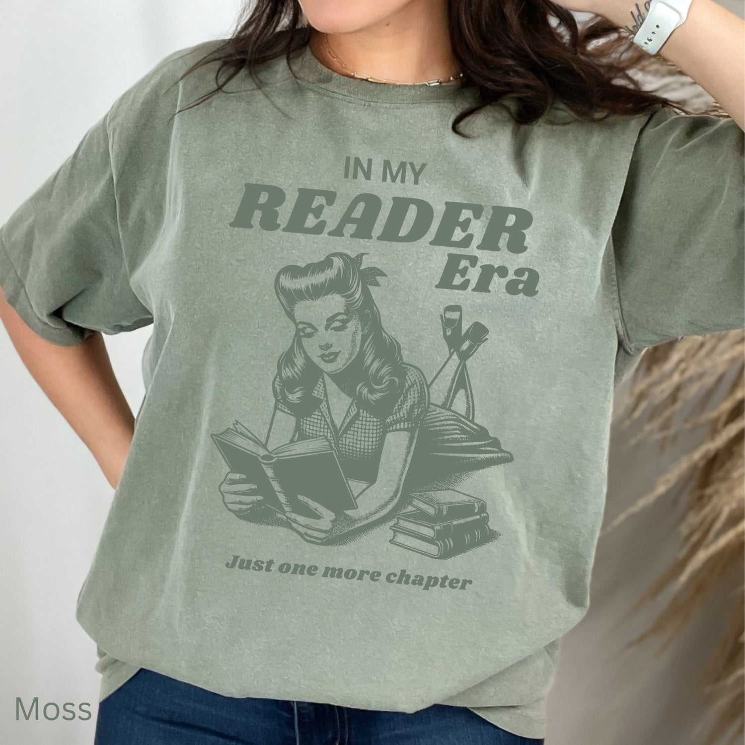 A woman wearing a Green Moss Comfort Colors tshirt, it has a book lover design on it that says "in my reader era" with a graphic of a woman reading a book.