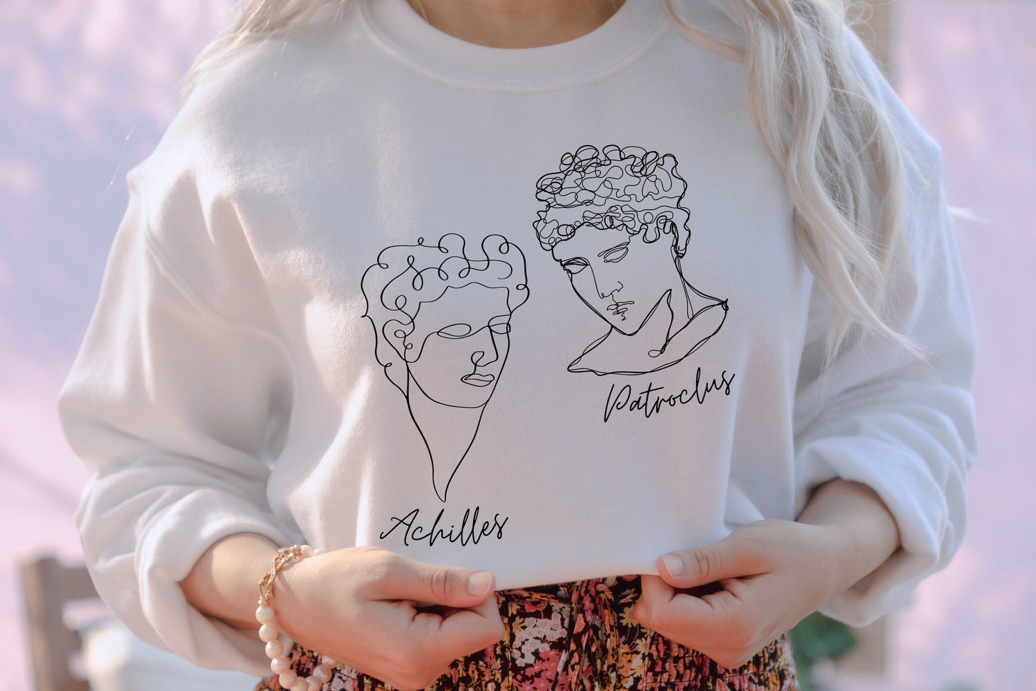 A woman wearing a white sweatshirt that has a graphic of Achilles & Patroclus as line art. From Starlit Prose Song of Achilles collection.
