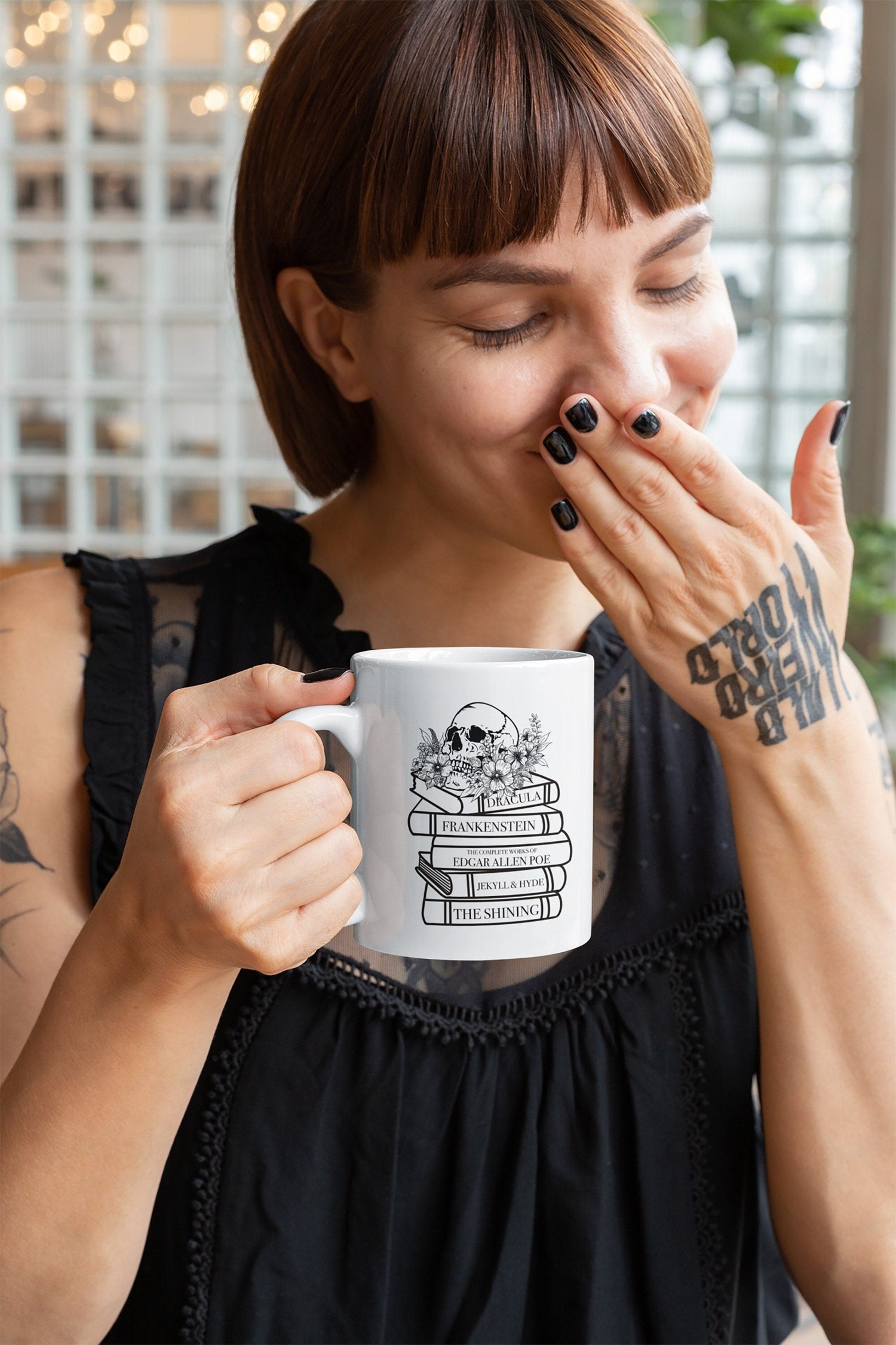 A woman holding a white mug that has a stack of books written by horror authors, for example Stephen King. It also has some flowers and a skull graphic on the mug too.