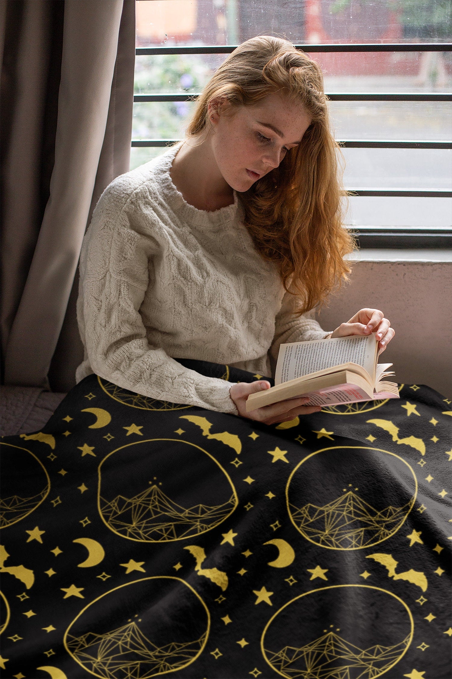 A woman reading, with a cozy blanket. The blanket has a pattern that is inspired by ACOTAR, it has a night court, stars, mountains and bats.