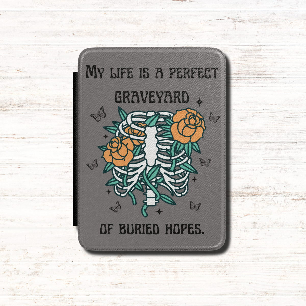 This dark literary Kindle case features the haunting quote "My life is a perfect graveyard of buried hopes" from Anne of Green Gables, paired with an illustrated skeletal ribcage from which delicate flowers and twisting vines emerge. It has a dark grey background. Designed by Starlit Prose Bookish Merch Australia.