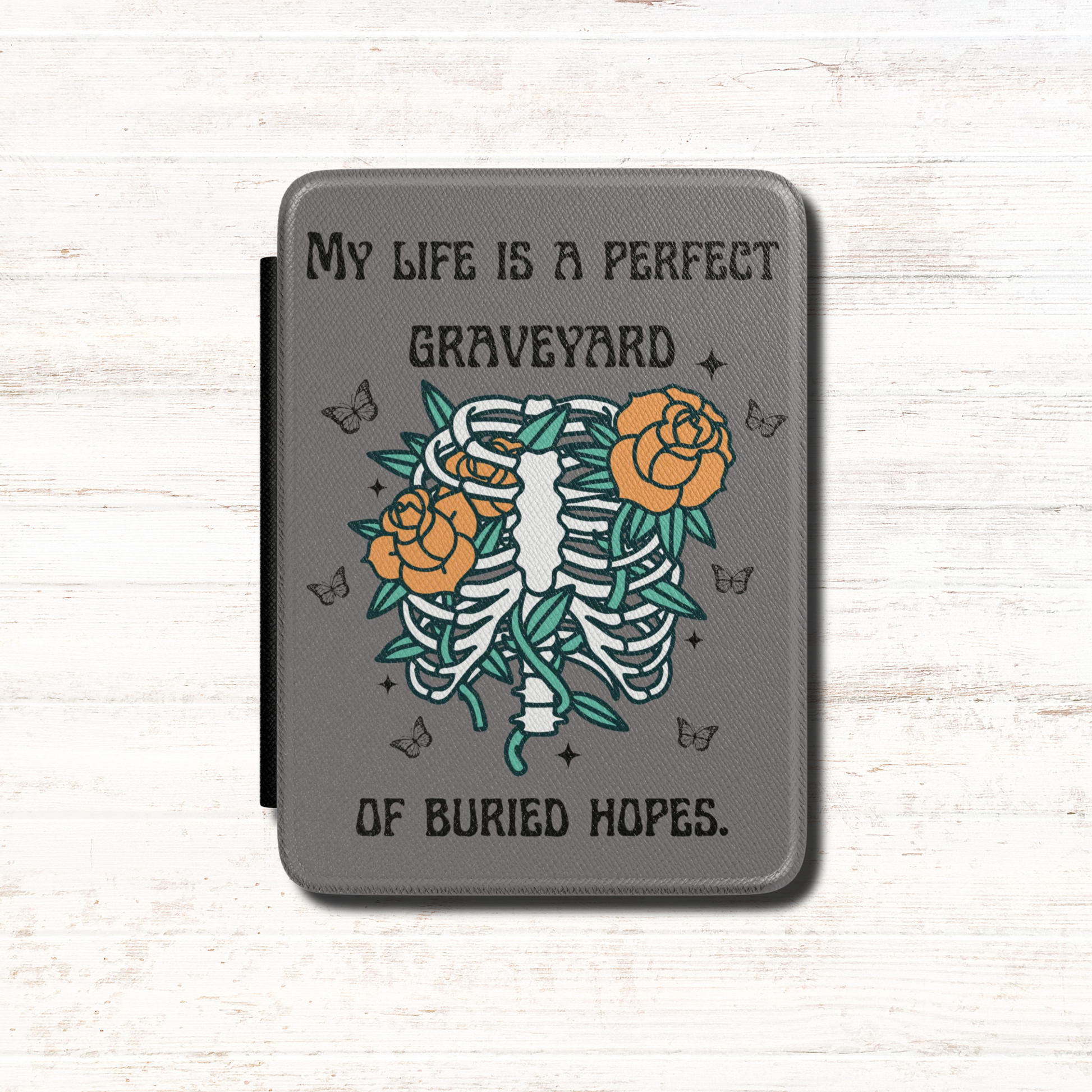 This dark literary Kindle case features the haunting quote "My life is a perfect graveyard of buried hopes" from Anne of Green Gables, paired with an illustrated skeletal ribcage from which delicate flowers and twisting vines emerge. It has a dark grey background. Designed by Starlit Prose Bookish Merch Australia.