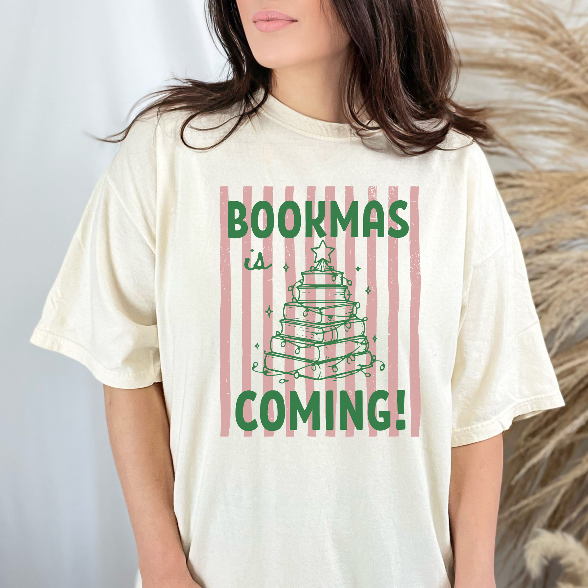 A woman wearing a Ivory Comfort Colors t-shirt, it has a pink distressed stripe pattern in the background, with bold green lettering that says "Bookmas is Coming!", it includes a green doodle illustration of a Christmas Tree made of books. Designed by Starlit Prose Bookish Merch Australia.