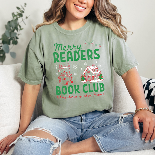 A woman wearing a Bay Green Comfort Colors t-shirt that reads 'Merry Readers Book Club' in bold green text, with cute doodle illustrations of a reading reindeer and a a cozy gingerbread house. Underneath is cursive red text that says ' Where stories spark joy forever". Designed by Starlit Prose Bookish Merch Australia.