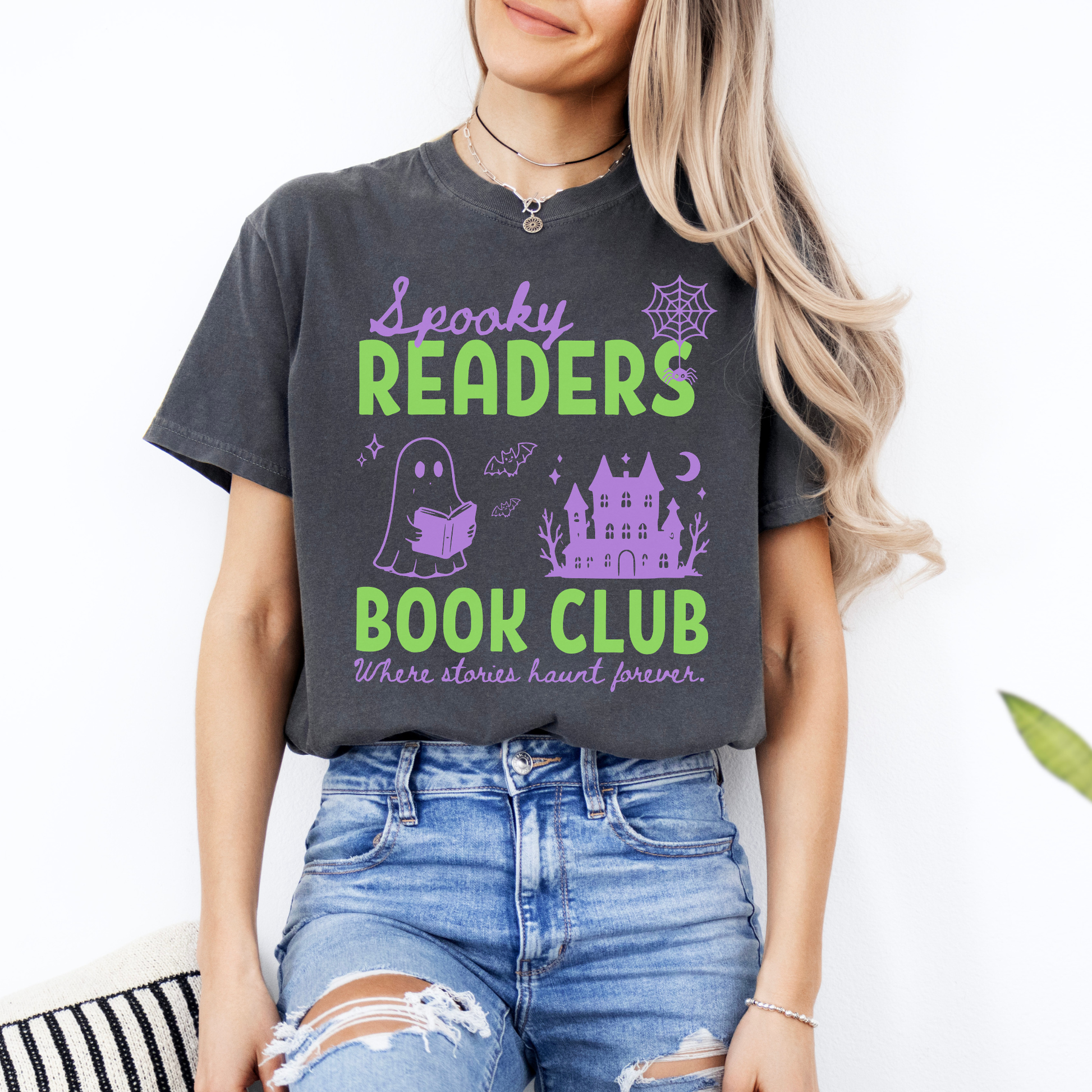  A woman wearing a Pepper Comfort colors graphic t-shirt with 'Spooky Readers Book Club' printed in green and purple, with Purple graphics including a cute ghost reading a book, a spider in its web & a haunted mansion paired with distressed blue jeans. Designed by Starlit Prose Bookish Merch Australia