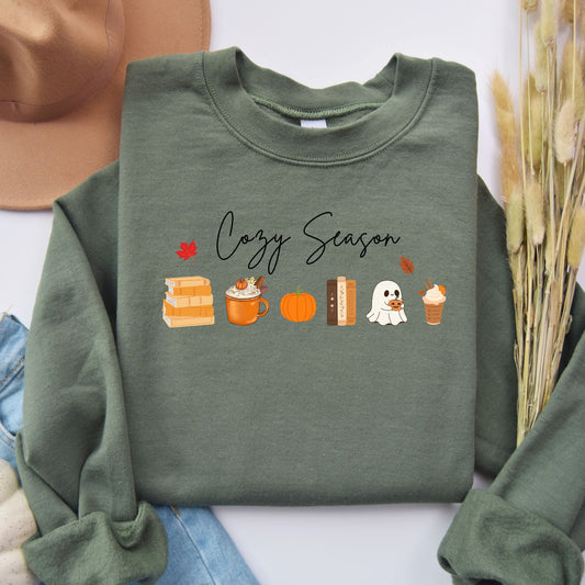 A Flatlay of a Green sweatshirt that says "Cozy Season", it includes cute orange Fall Autumn graphics like a stack of books, pumpkin spice latte, pumpkins, a ghost and leaves. Starlit Prose Bookish Merch Australia