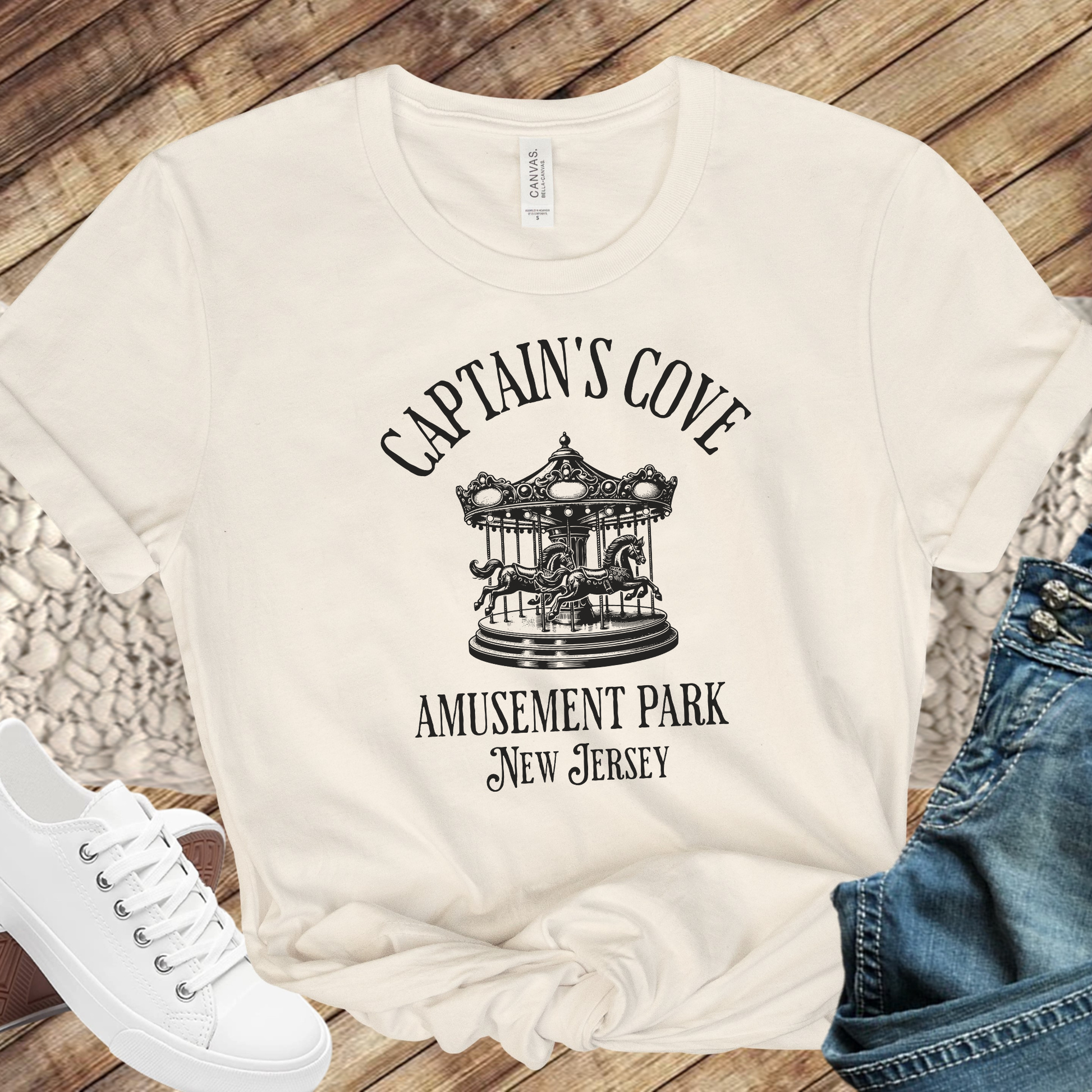Natural t-shirt with 'Captain's Cove Amusement Park New Jersey' design on a wooden surface with jeans and sneakers.