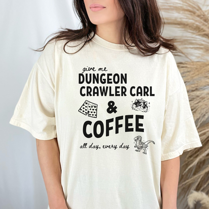 A woman wearing an oversized Ivory graphic Comfort colors T-shirt with black text reading 'give me DUNGEON CRAWLER CARL & COFFEE all day, every day' and small illustrations - including Carl's pajama shorts, Princess Donut & Mongo. Designed by Starlit Prose, Bookish Merch Australia