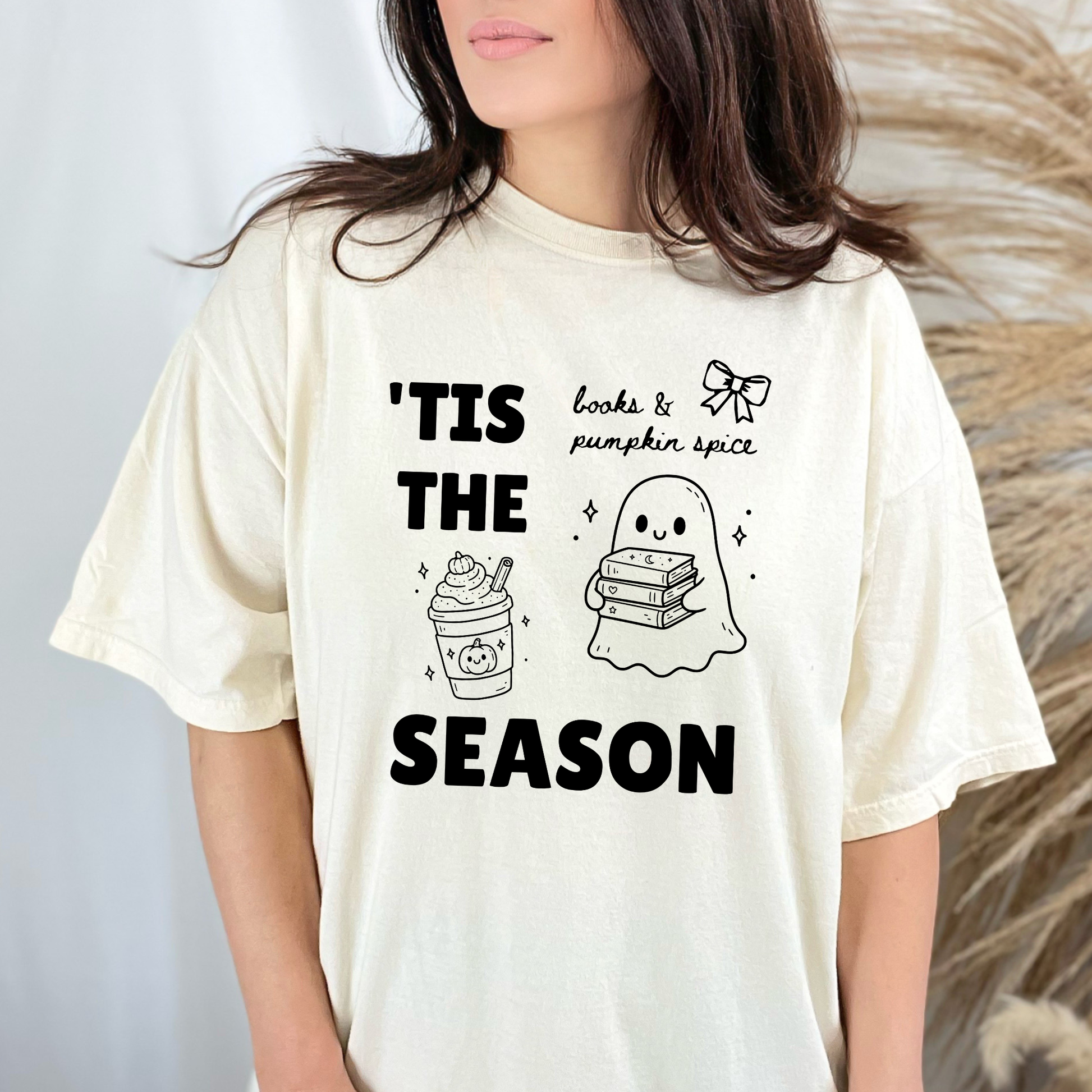 A woman wearing a Ivory graphic t-shirt with 'TIS THE SEASON' in bold font with cute Halloween-themed illustrations including a ghost holding a stack of books, a cute pumpkin spiced latte and a coquette bow with the phrase "books & pumpkin spice". Designed by Starlit Prose Bookish Merch Australia.