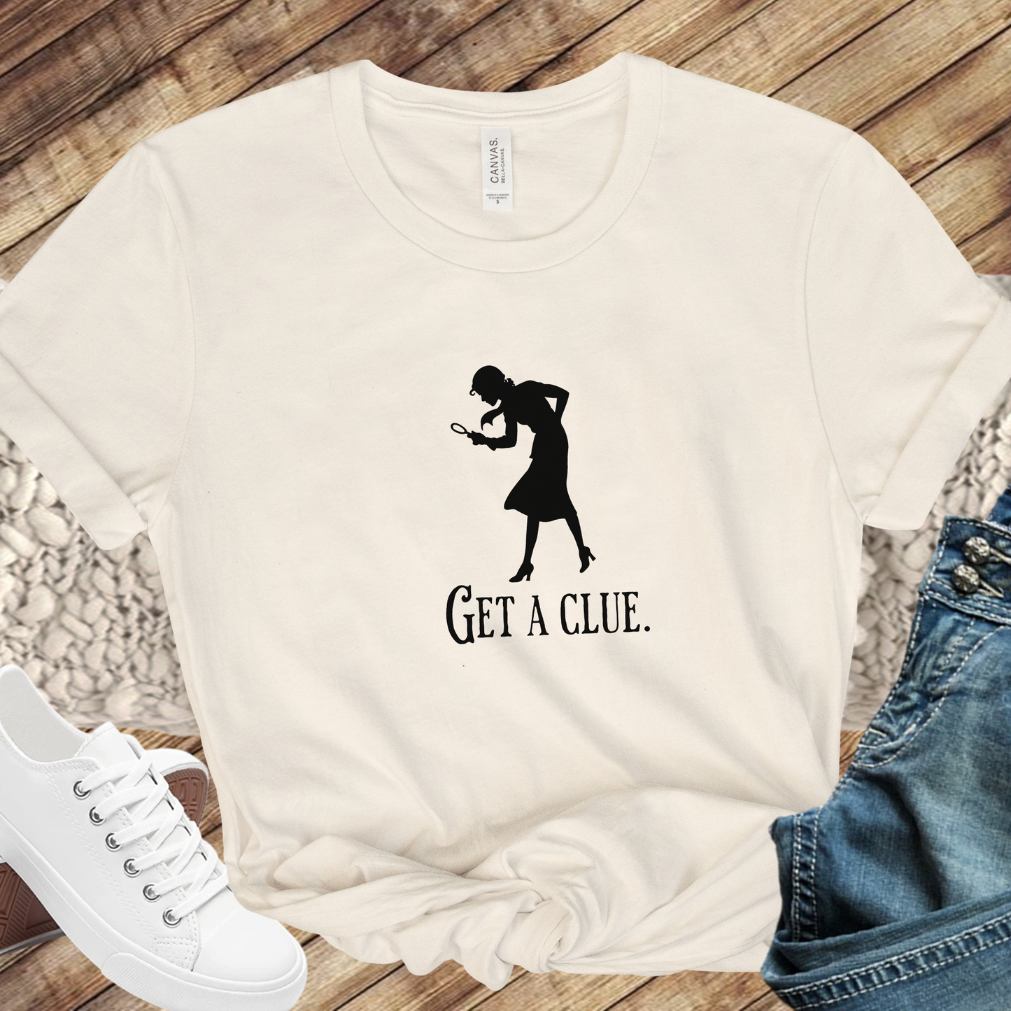 Beige t-shirt with a black silhouette and 'GET A CLUE.' text on a wooden surface with jeans and sneakers.