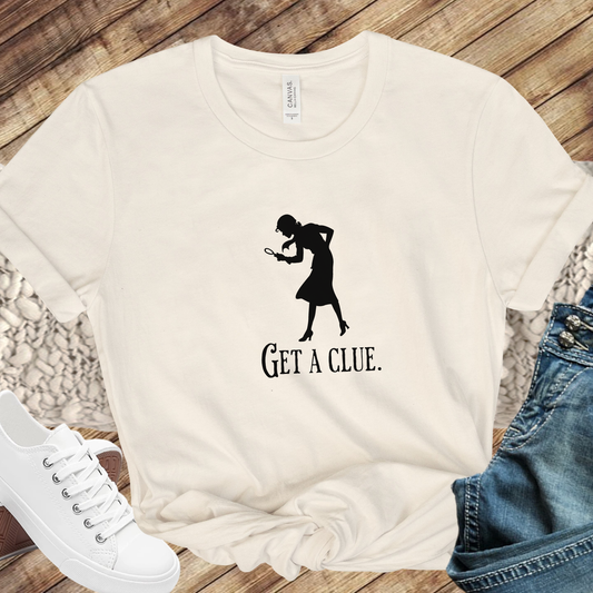 Beige t-shirt with a black silhouette and 'GET A CLUE.' text on a wooden surface with jeans and sneakers.