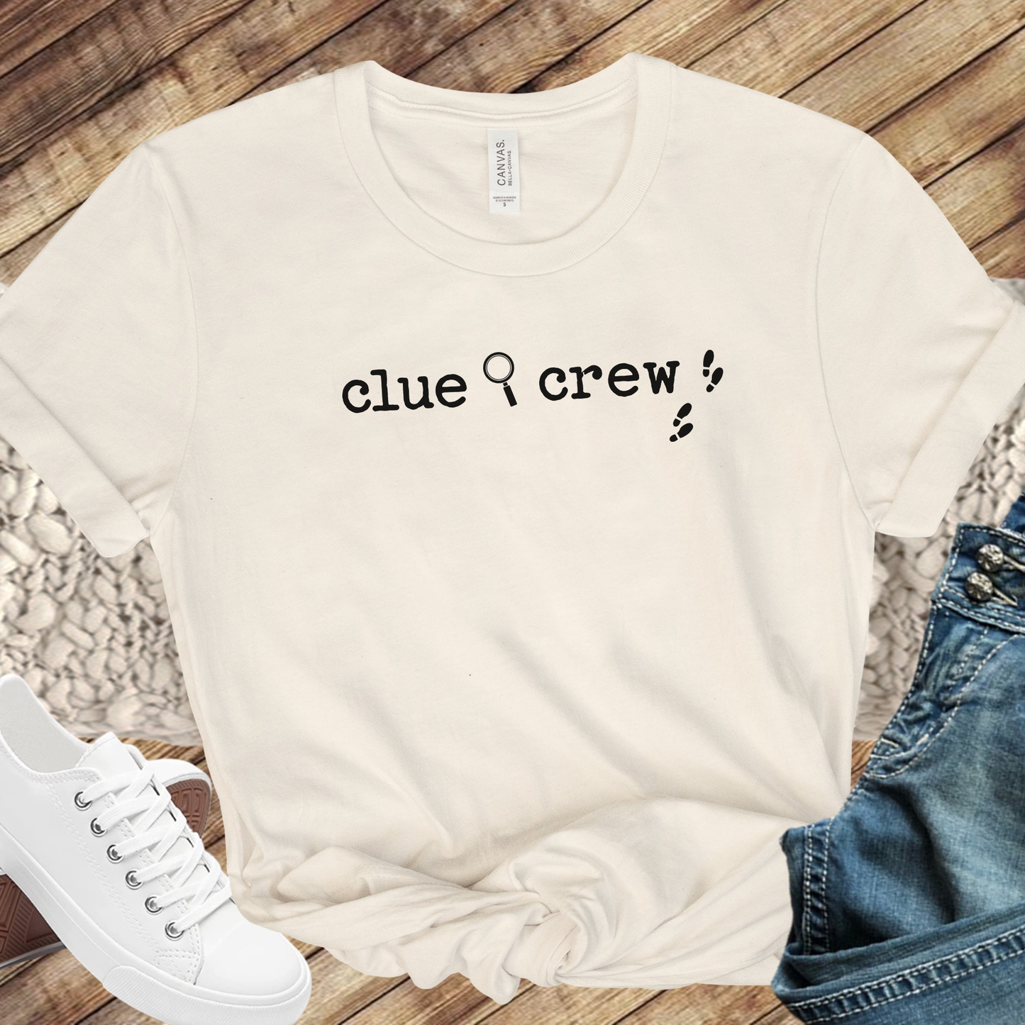 Natural t-shirt with 'clue crew' text on a wooden surface with jeans and white sneakers.