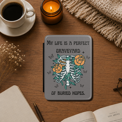 This dark literary Kindle case features the haunting quote "My life is a perfect graveyard of buried hopes" from Anne of Green Gables, paired with an illustrated skeletal ribcage from which delicate flowers and twisting vines emerge. It has a dark grey background. Designed by Starlit Prose Bookish Merch Australia.