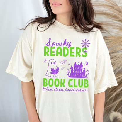  A woman wearing a Ivory Comfort colors graphic t-shirt with 'Spooky Readers Book Club' printed in green and purple, with Purple graphics including a cute ghost reading a book, a spider in its web & a haunted mansion paired with distressed blue jeans. Designed by Starlit Prose Bookish Merch Australia