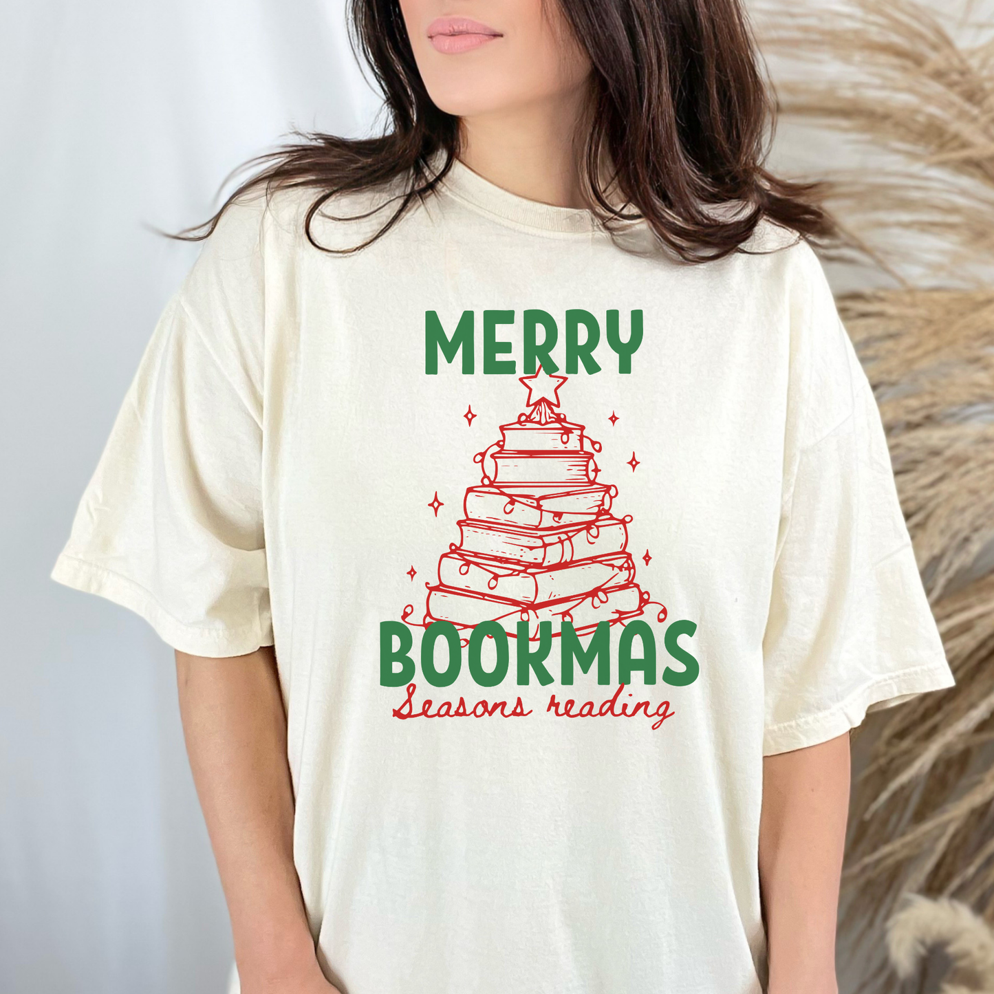 A woman wearing a Ivory Comfort Colors t-shirt with bold text that says "Merry Bookmas" in green, and "Seasons Reading" in red cursive font, includes a doodle illustration of a Christmas tree made of books. Designed by Starlit Prose Bookish Merch Australia.