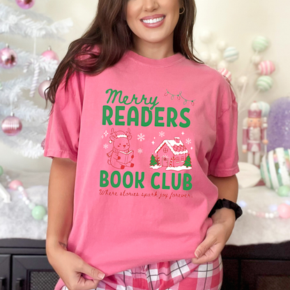 A woman wearing a Crunchberry Pink Comfort Colors t-shirt that reads 'Merry Readers Book Club' in bold green text, with cute doodle illustrations of a reading reindeer and a a cozy gingerbread house. Underneath is cursive red text that says ' Where stories spark joy forever". Designed by Starlit Prose Bookish Merch Australia.