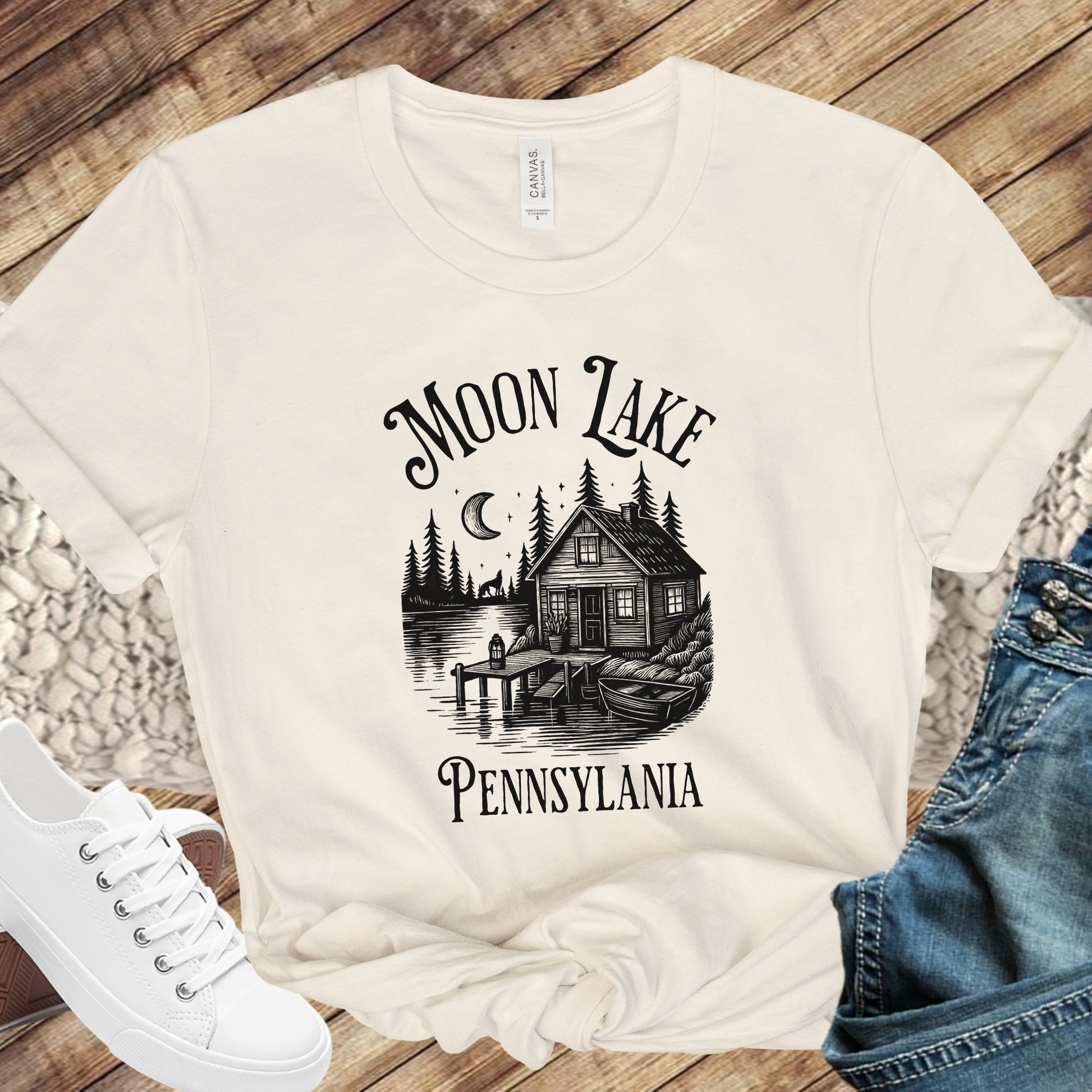 Beige t-shirt with 'Moon Lake Pennsylvania' graphic design on a wooden surface with shoes and jeans.