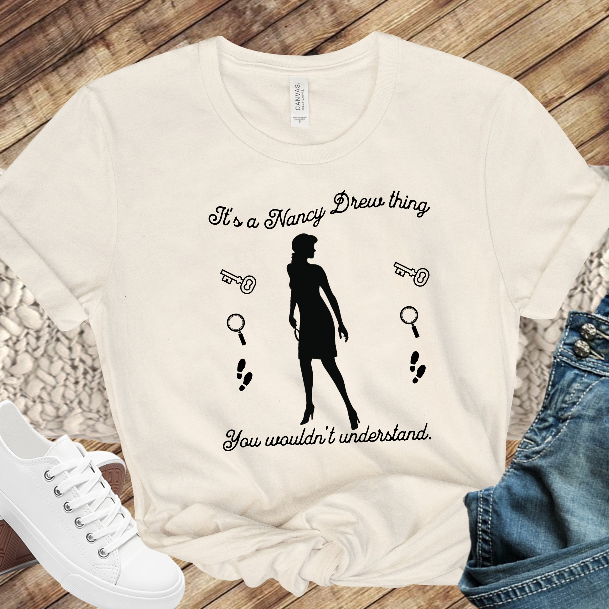 Natural tshirt with text that says "It's a Nancy Drew thing, you wouldn't understand" with graphic elements like a key, magnifying glass and footsteps.