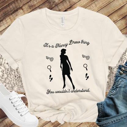 Natural tshirt with text that says "It's a Nancy Drew thing, you wouldn't understand" with graphic elements like a key, magnifying glass and footsteps.