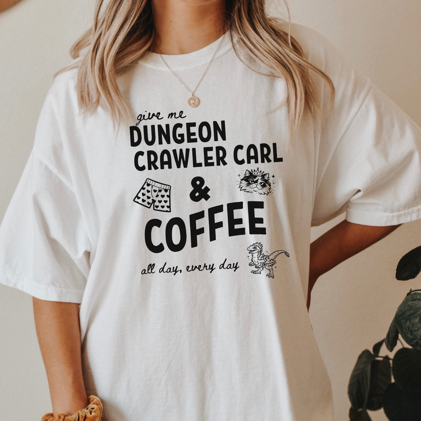 A woman wearing an oversized white graphic Comfort colors T-shirt with black text reading 'give me DUNGEON CRAWLER CARL & COFFEE all day, every day' and small illustrations - including Carl's pajama shorts, Princess Donut & Mongo. Designed by Starlit Prose, Bookish Merch Australia