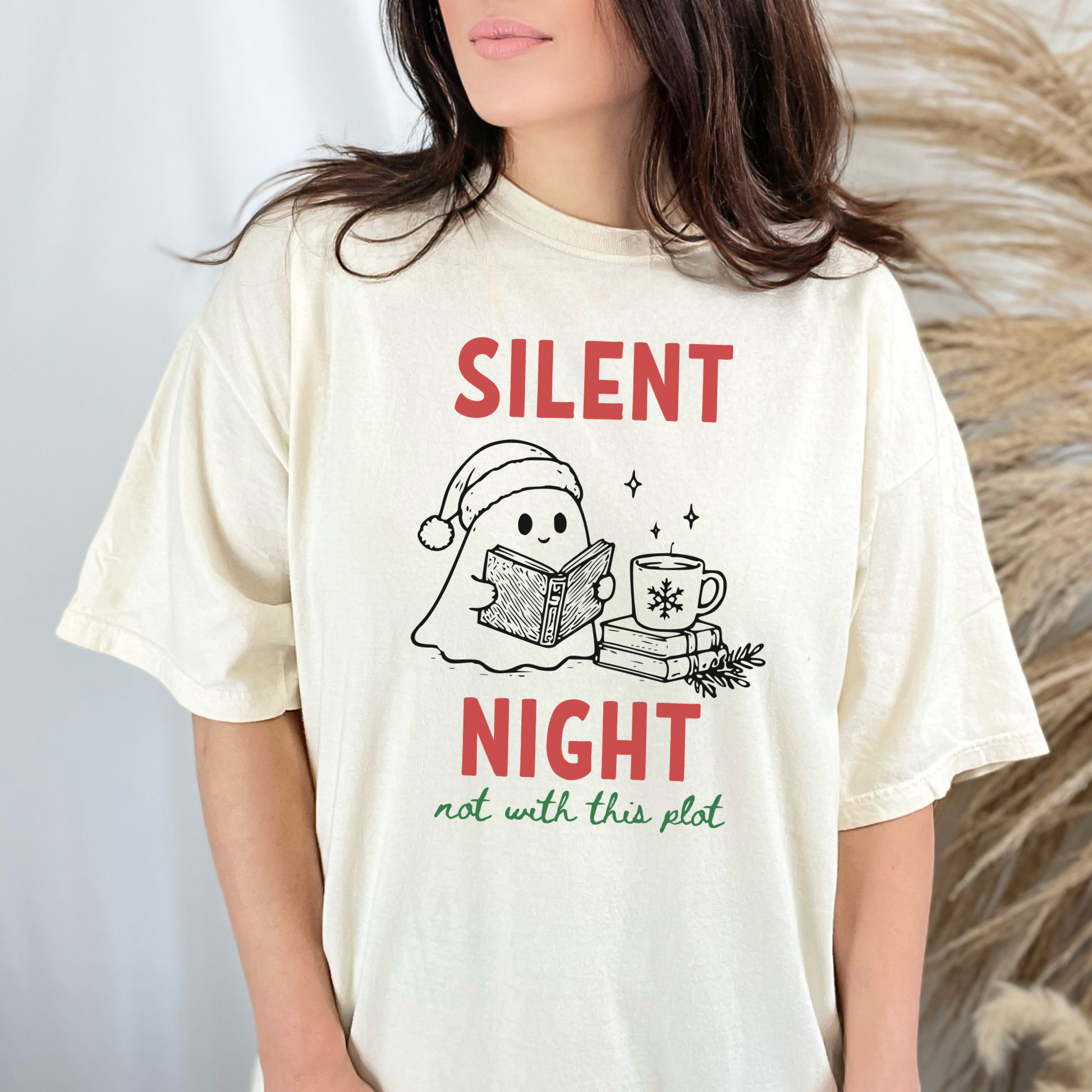 A woman wearing an Ivory Comfort Colors t-shirt with a cute doodle illustration of a ghost reading a book, with a cup of coffee stacked on some books. There's bold text that says  'SILENT NIGHT" in red, with green cursive that says " not with this plot". Designed by Starlit Prose Bookish Merch Australia.