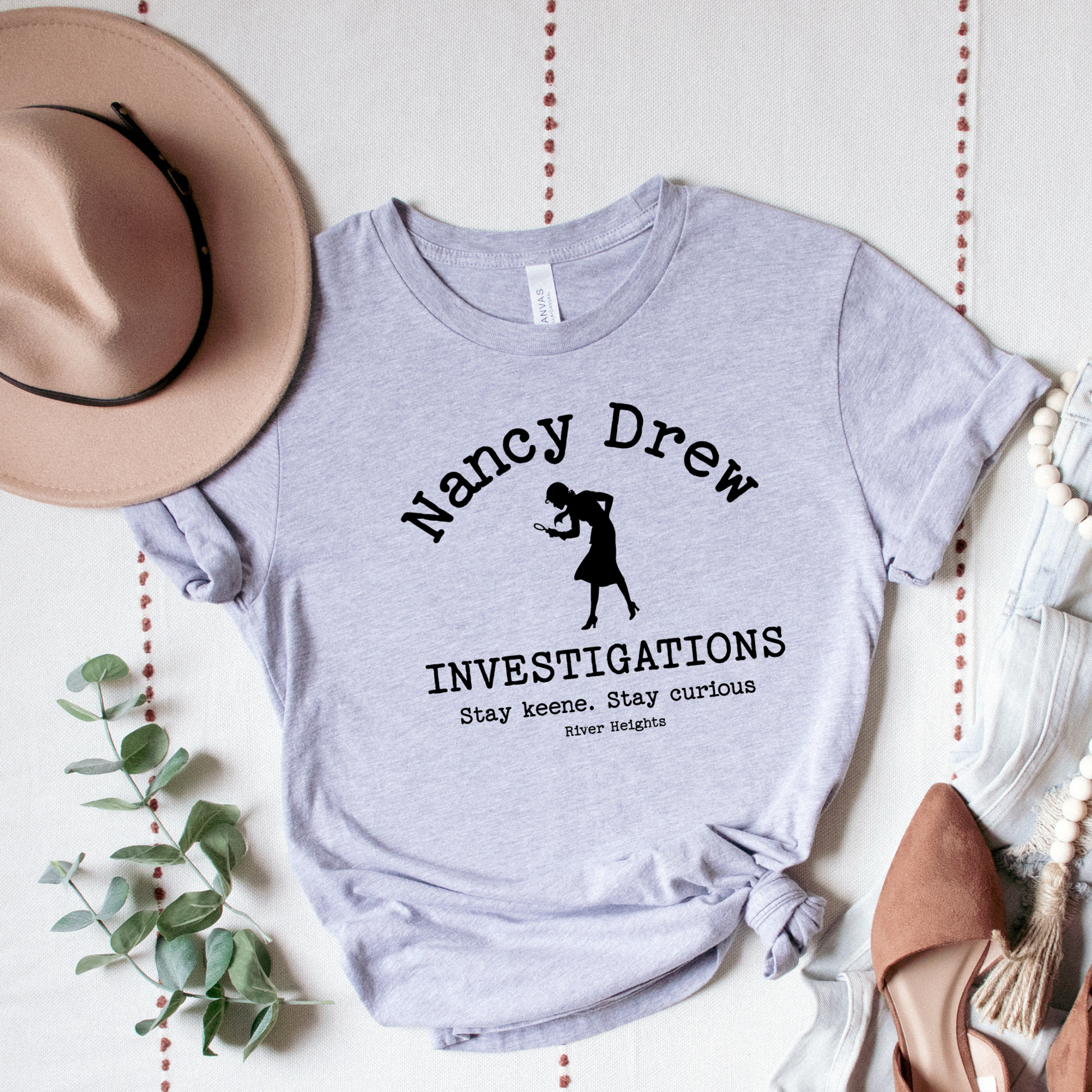 Gray t-shirt with 'Nancy Drew Investigations' design on a white surface with decorative elements.