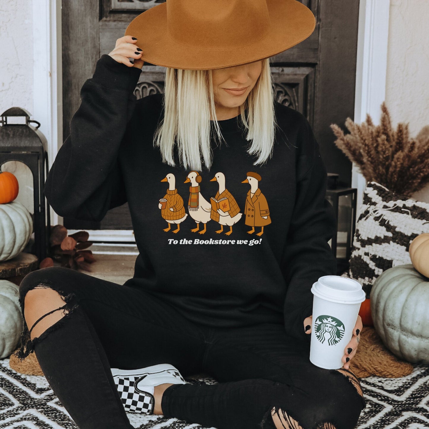 Person wearing a black sweatshirt with a graphic design of a group of ducks going to a bookstore, holding a Starbucks cup, in a cozy indoor setting. Starlit Prose Bookish Merch Australia