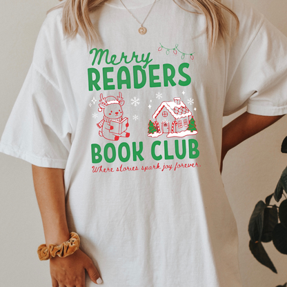 A woman wearing a White Comfort Colors t-shirt that reads 'Merry Readers Book Club' in bold green text, with cute doodle illustrations of a reading reindeer and a a cozy gingerbread house. Underneath is cursive red text that says ' Where stories spark joy forever". Designed by Starlit Prose Bookish Merch Australia.