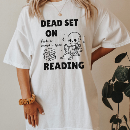 A woman wearing a White Comfort Colors  t-shirt with a black print that reads 'DEAD SET ON READING' alongside a skeleton reading books and a stack of books. It also says books & pumpkin spice in script font. Designed by Starlit Prose Bookish Merch Australia