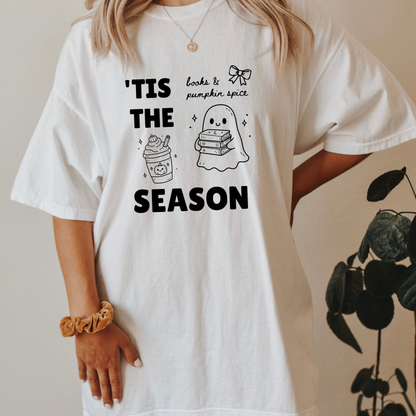 A woman wearing a White graphic t-shirt with 'TIS THE SEASON' in bold font with cute Halloween-themed illustrations including a ghost holding a stack of books, a cute pumpkin spiced latte and a coquette bow with the phrase "books & pumpkin spice". Designed by Starlit Prose Bookish Merch Australia.
