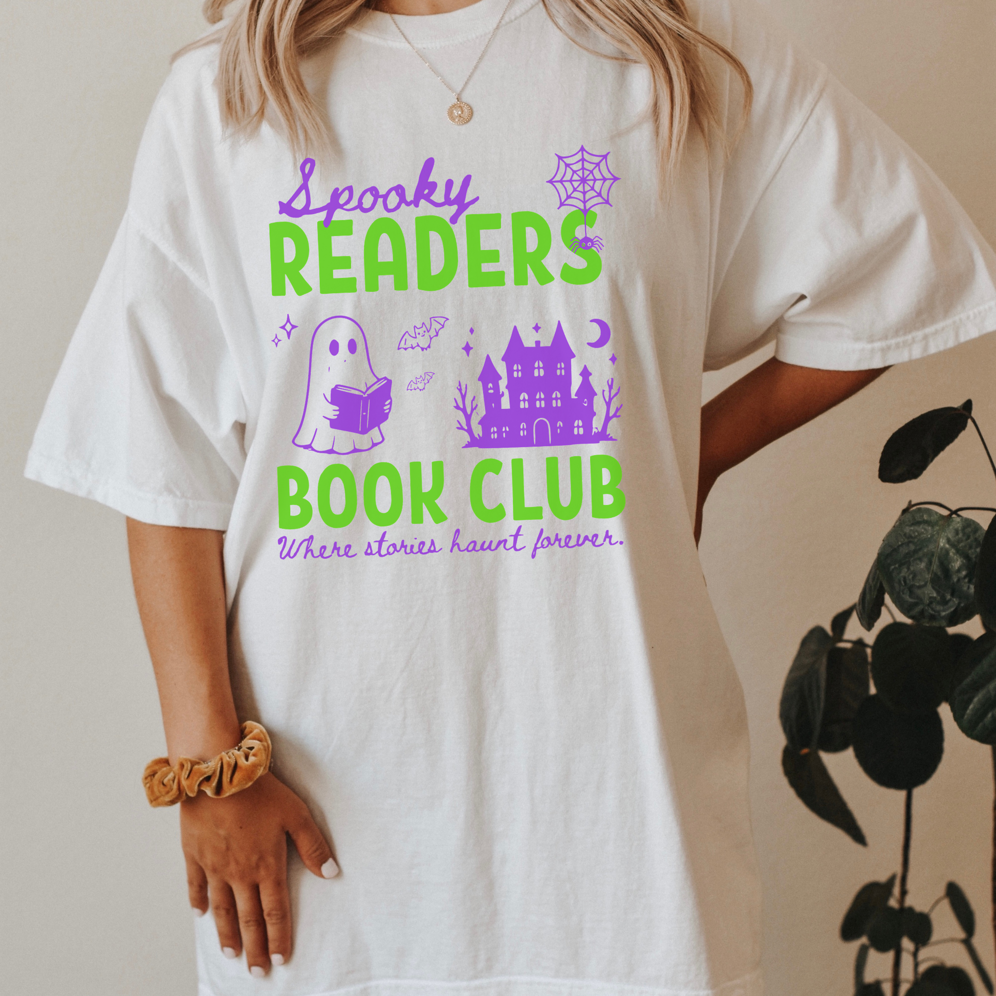  A woman wearing a White Comfort colors graphic t-shirt with 'Spooky Readers Book Club' printed in green and purple, with Purple graphics including a cute ghost reading a book, a spider in its web & a haunted mansion paired with distressed blue jeans. Designed by Starlit Prose Bookish Merch Australia