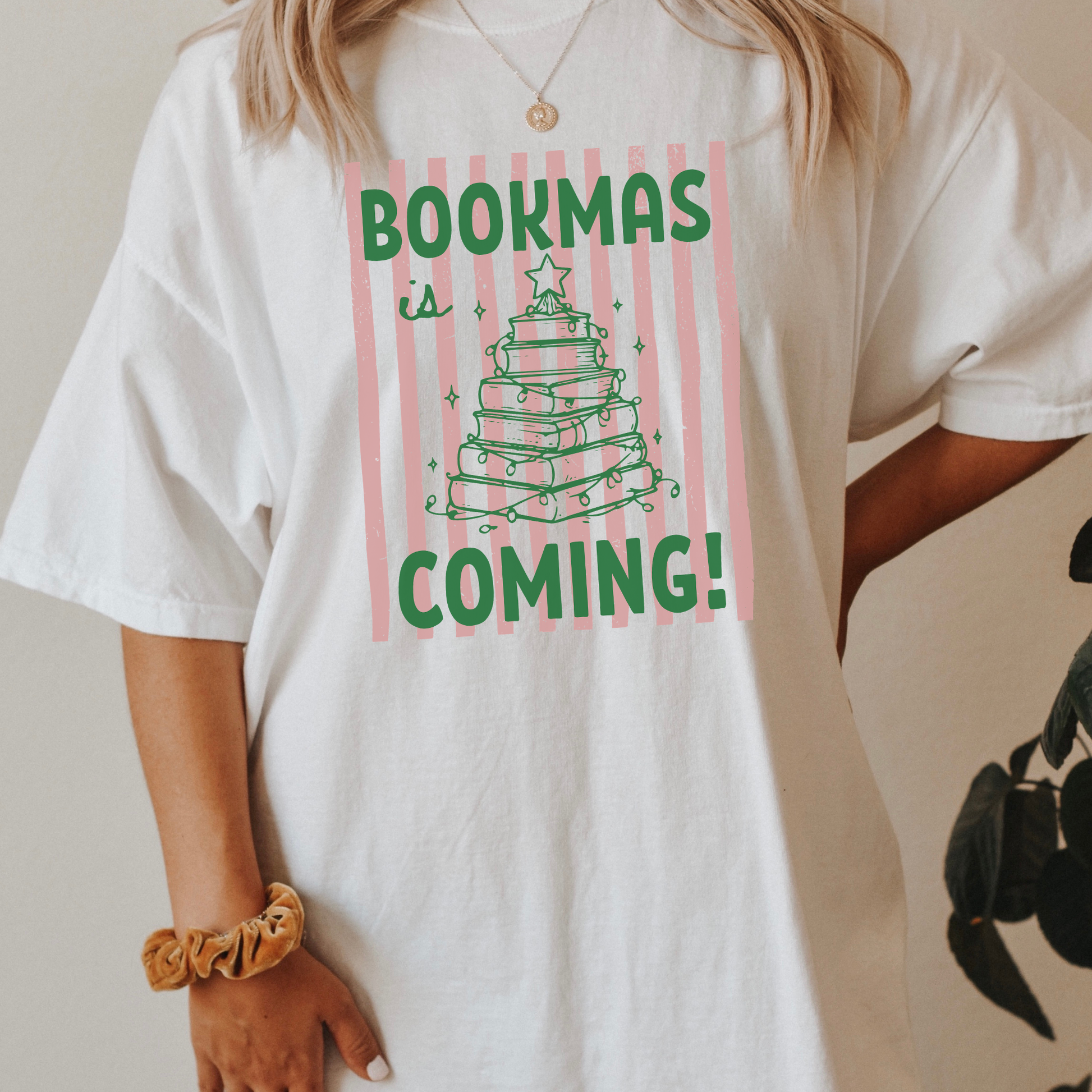 A woman wearing a White Comfort Colors t-shirt, it has a pink distressed stripe pattern in the background, with bold green lettering that says "Bookmas is Coming!", it includes a green doodle illustration of a Christmas Tree made of books. Designed by Starlit Prose Bookish Merch Australia.