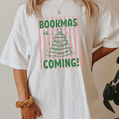 A woman wearing a White Comfort Colors t-shirt, it has a pink distressed stripe pattern in the background, with bold green lettering that says "Bookmas is Coming!", it includes a green doodle illustration of a Christmas Tree made of books. Designed by Starlit Prose Bookish Merch Australia.