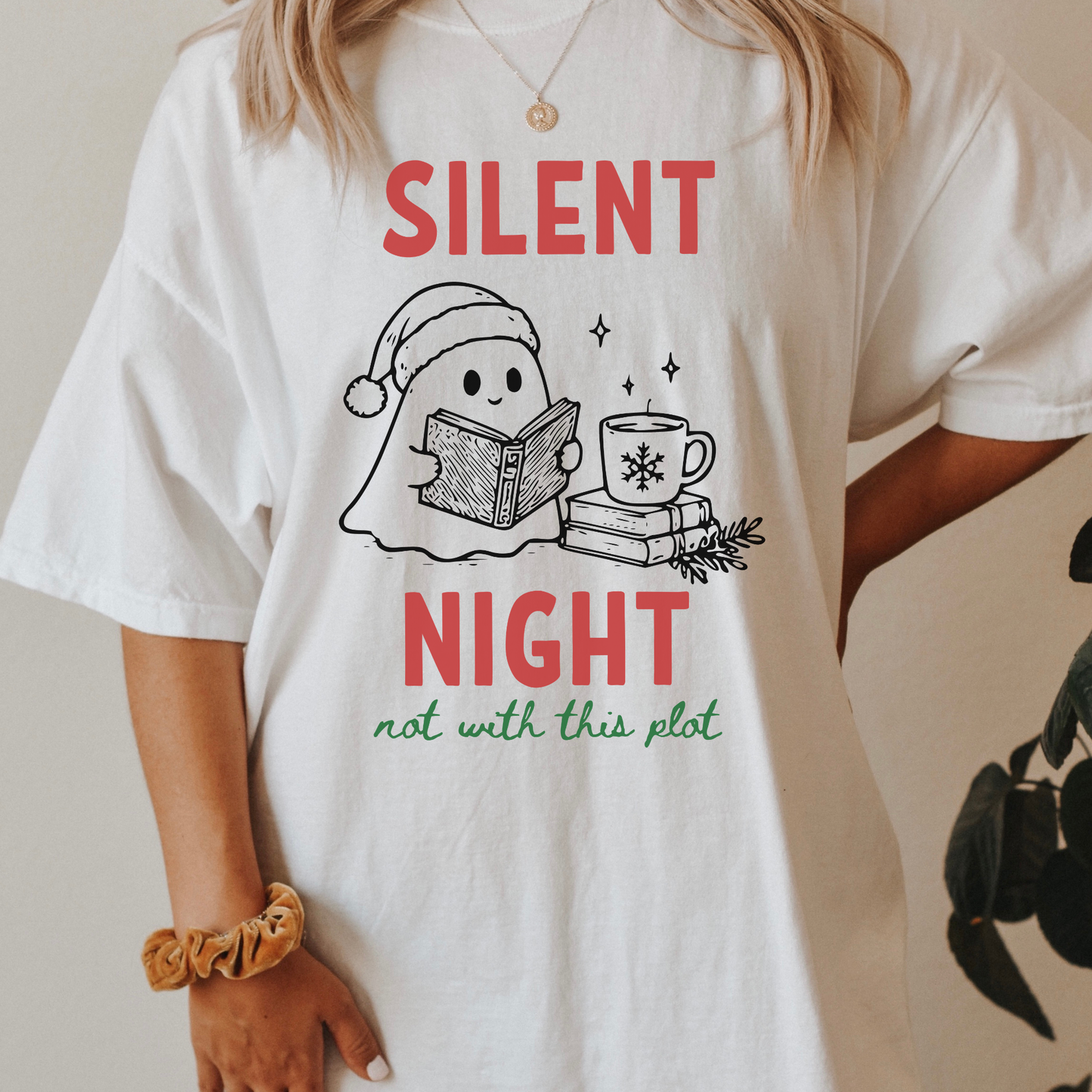 A woman wearing a white Comfort Colors t-shirt with a cute doodle illustration of a ghost reading a book, with a cup of coffee stacked on some books. There's bold text that says  'SILENT NIGHT" in red, with green cursive that says " not with this plot". Designed by Starlit Prose Bookish Merch Australia.