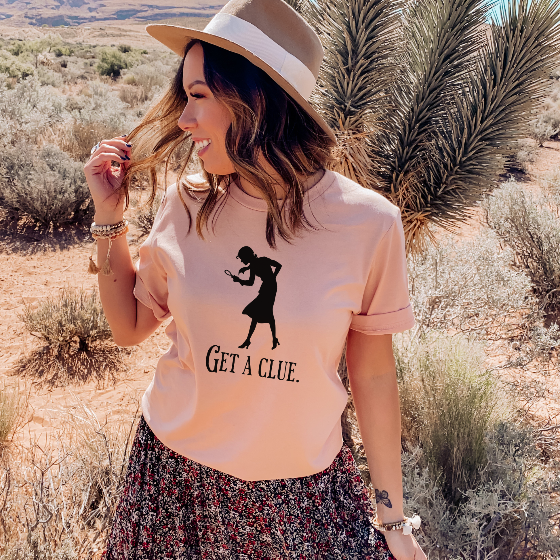 Woman in a desert setting wearing a heather peach t-shirt with a graphic and text.