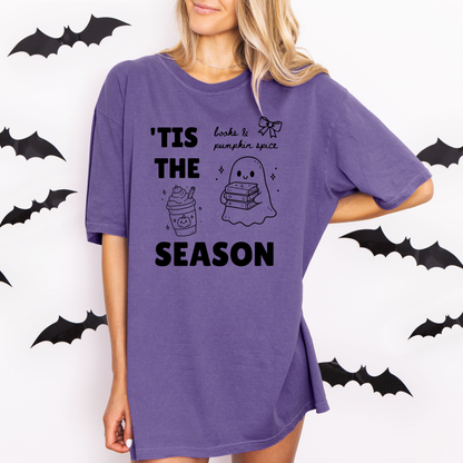 A woman wearing a Grape graphic t-shirt with 'TIS THE SEASON' in bold font with cute Halloween-themed illustrations including a ghost holding a stack of books, a cute pumpkin spiced latte and a coquette bow with the phrase "books & pumpkin spice". Designed by Starlit Prose Bookish Merch Australia.